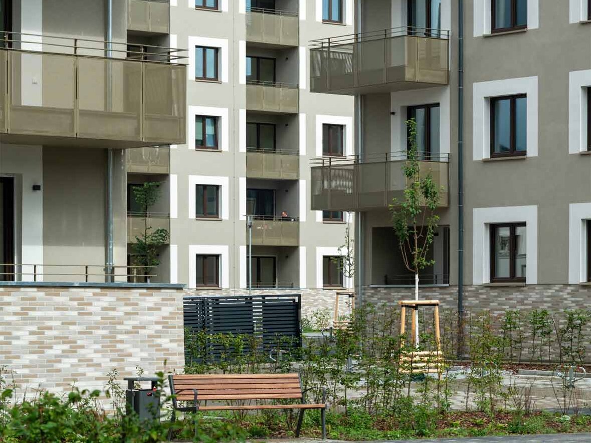 Außenansicht degewo-Neubau Falkenstraße Adlerstraße: Hofseite mit Sträuchern im Beet, Mülleimer und Sitzbank im Vordergrund.