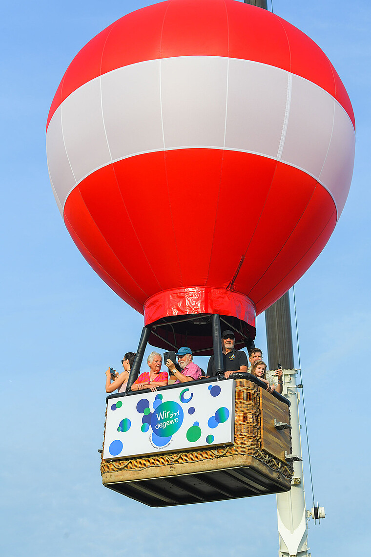 Mehrere Personen befinden sich in einem Heißluftballon. Dieser wird von einem Baukran in den Himmel hinauf gezogen. 