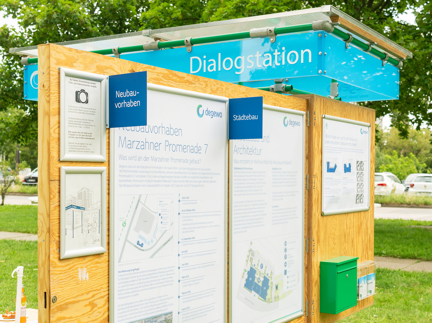 Die degewo-Dialogstation ist im aufgeklappten Zustand. An einer Holzwand hängen mehrere große Plakate, daneben ein Briefkasten. Auf der Rückseite ein kleiner Tisch mit Hockern.