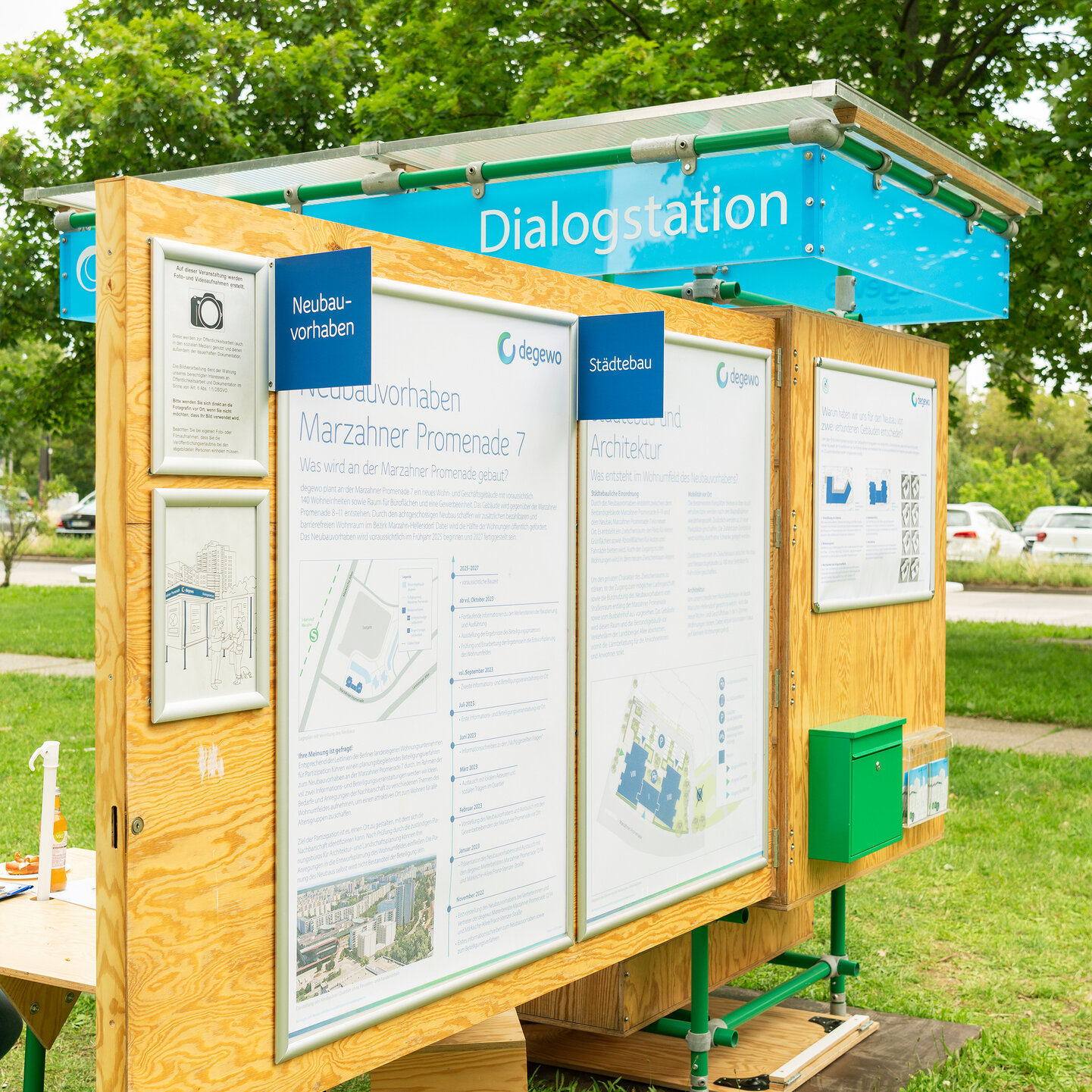Die degewo-Dialogstation ist im aufgeklappten Zustand. An einer Holzwand hängen mehrere große Plakate, daneben ein Briefkasten. Auf der Rückseite ein kleiner Tisch mit Hockern.