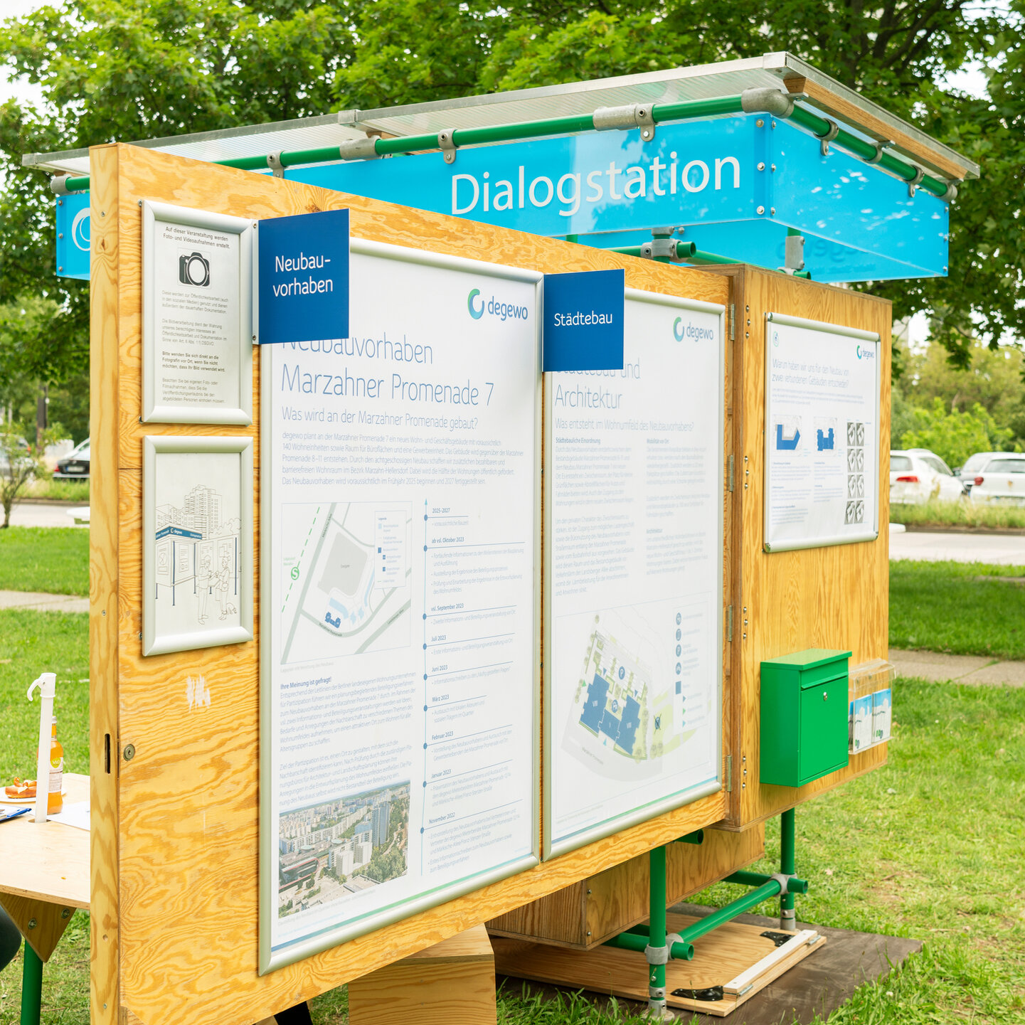 Die degewo-Dialogstation ist im aufgeklappten Zustand. An einer Holzwand hängen mehrere große Plakate, daneben ein Briefkasten. Auf der Rückseite ein kleiner Tisch mit Hockern.