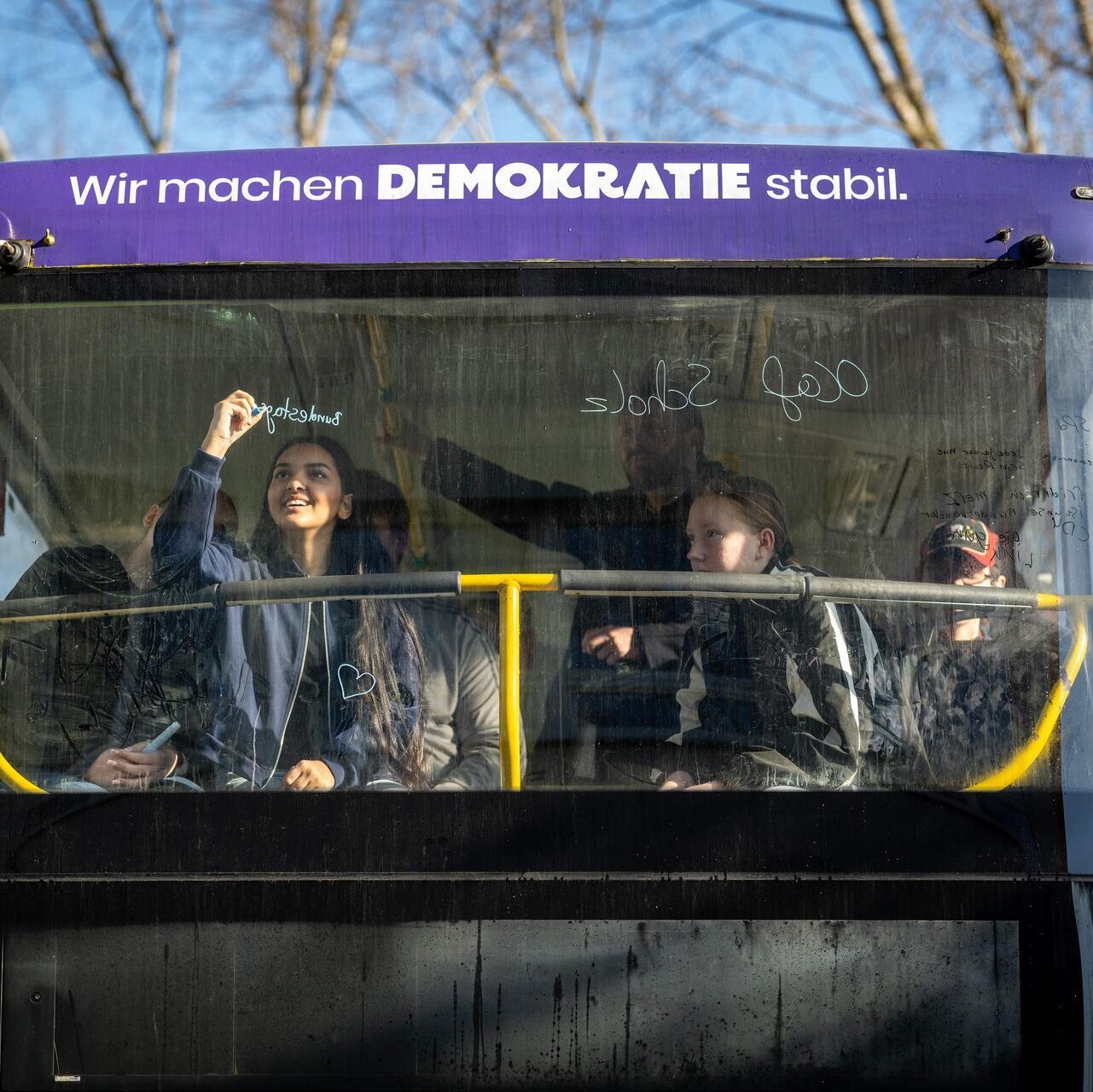 Blick von Außen auf die Frontscheibe eines Doppeldecker-Busses mit der Aufschrift: Wir machen Demokratie stabil. Hinter der Scheibe schreibt ein Mädchen etwas ans Fenster mit einem bunten Stift.