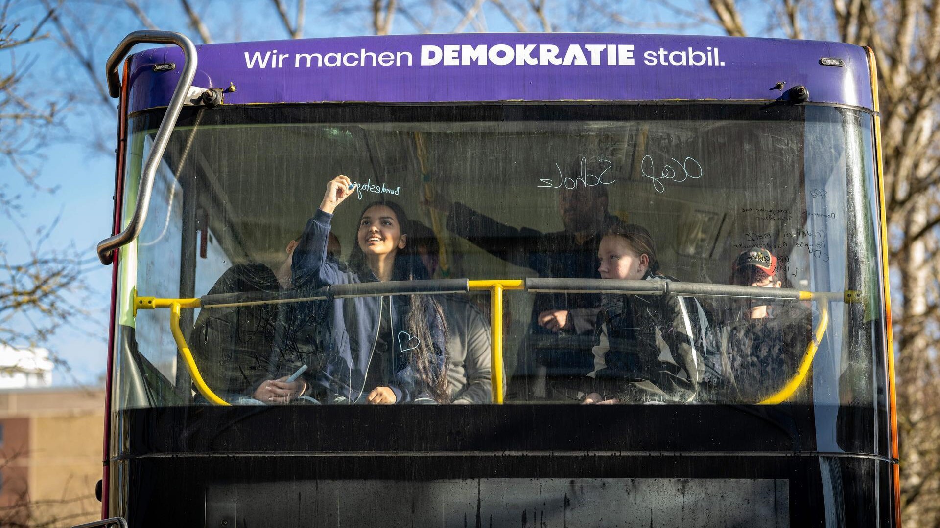 Blick von Außen auf die Frontscheibe eines Doppeldecker-Busses mit der Aufschrift: Wir machen Demokratie stabil. Hinter der Scheibe schreibt ein Mädchen etwas ans Fenster mit einem bunten Stift.