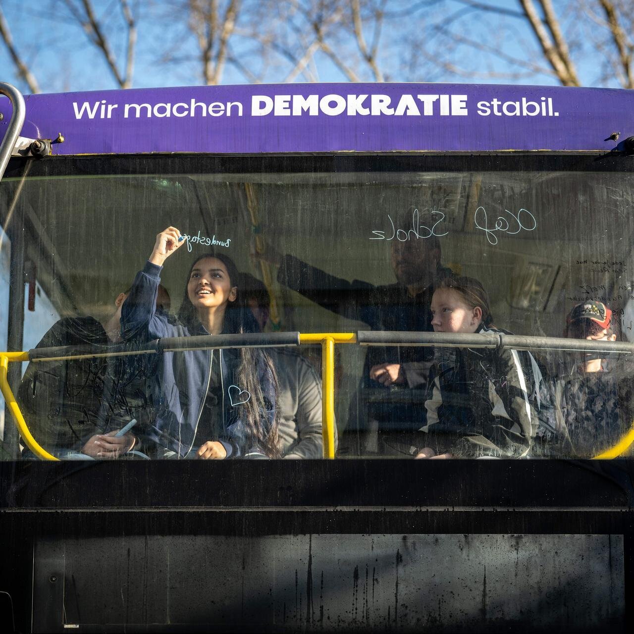 Blick von Außen auf die Frontscheibe eines Doppeldecker-Busses mit der Aufschrift: Wir machen Demokratie stabil. Hinter der Scheibe schreibt ein Mädchen etwas ans Fenster mit einem bunten Stift.
