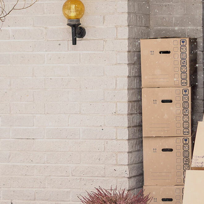 Das Foto zeigt einen Mann in blauer Jacke, wie er drei aufeinandergestapelte Kartons auf einer Sackkarre aus dem Haus schiebt. Es schneit.