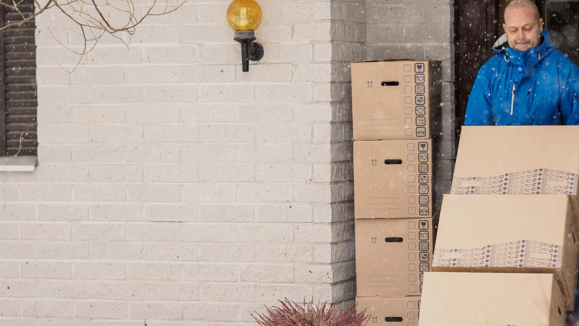 Das Foto zeigt einen Mann in blauer Jacke, wie er drei aufeinandergestapelte Kartons auf einer Sackkarre aus dem Haus schiebt. Es schneit.