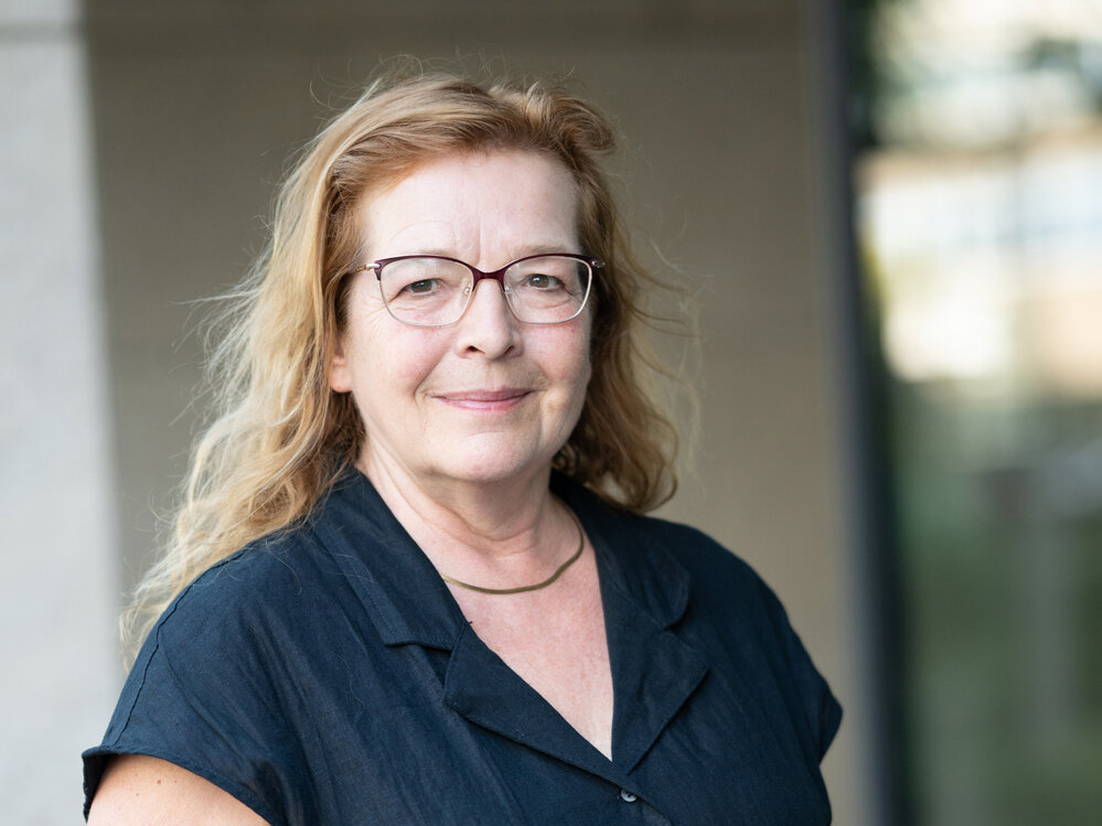 Porträt einer Frau mit heller, leicht gewellter Frisur und Brille, die in einer dunklen Bluse selbstbewusst vor einem Wohngebäude steht und freundlich in die Kamera lächelt.