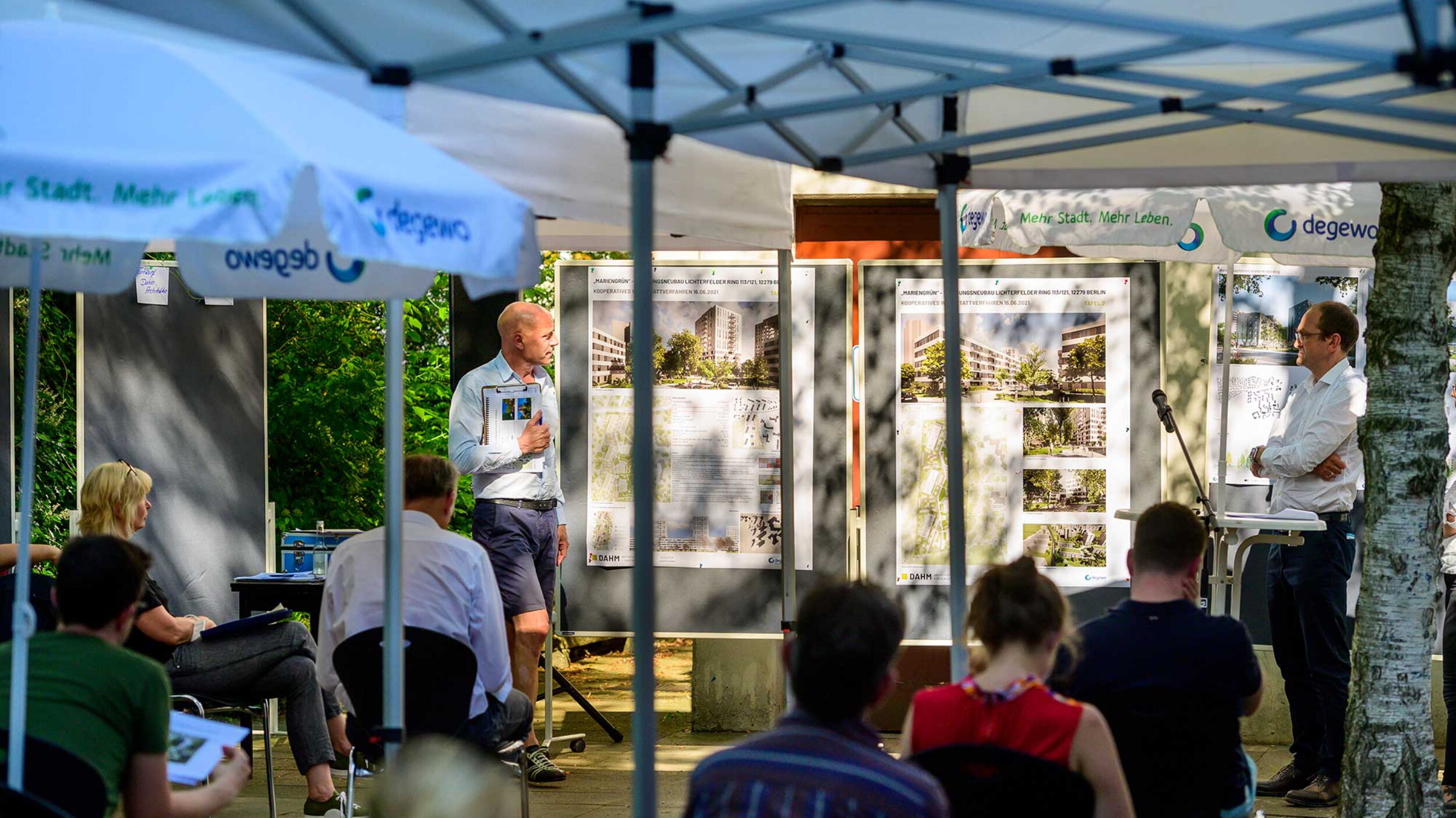 Mehrere Personen sitzen unter weißen Pavillons und hören zwei Personen zu. Im Hintergrund große Stelltafeln mit Plänen und Visualisierungen.