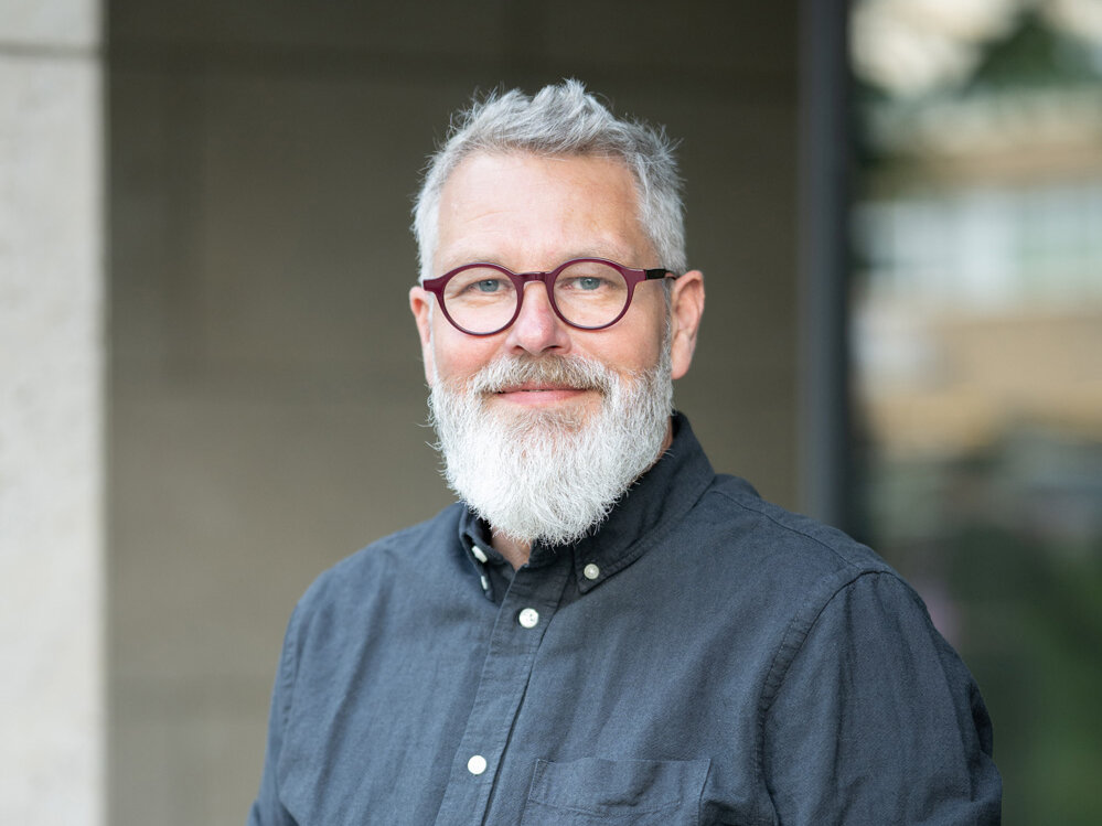 Mann mit grauem Bart und Brille trägt ein dunkles Hemd und steht im Freien vor einem Gebäude; er blickt freundlich in die Kamera, der Hintergrund ist weichgezeichnet.
