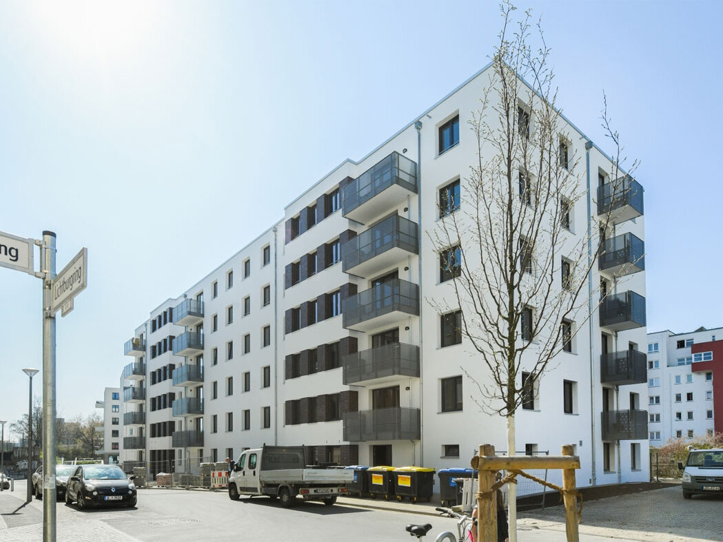 Straßeneckenansicht degewo-Neubau Lichtburgring mit Straßenschild und neu gepflanztem Baum im Vordergrund.