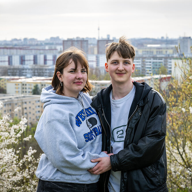 Ein junges Paar posiert auf einer Anhöhe. Im Hintergrund ist verschwommen das Berliner Stadtponarama zu erkennen.