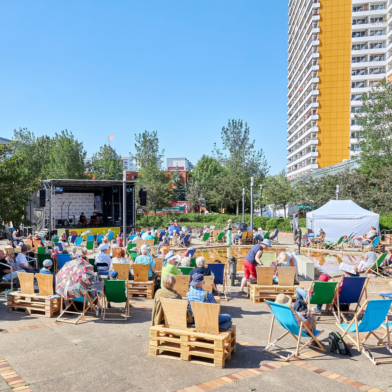 Ansicht des Helene-Weigel-Platzes vor blaunen Himmel und Sonnenschein. Es ist eine Bühne aufgebaut, Menschen sitzen auf Liegestühlen darum. Im Hintergrund ist ein Hochhaus zu sehen.