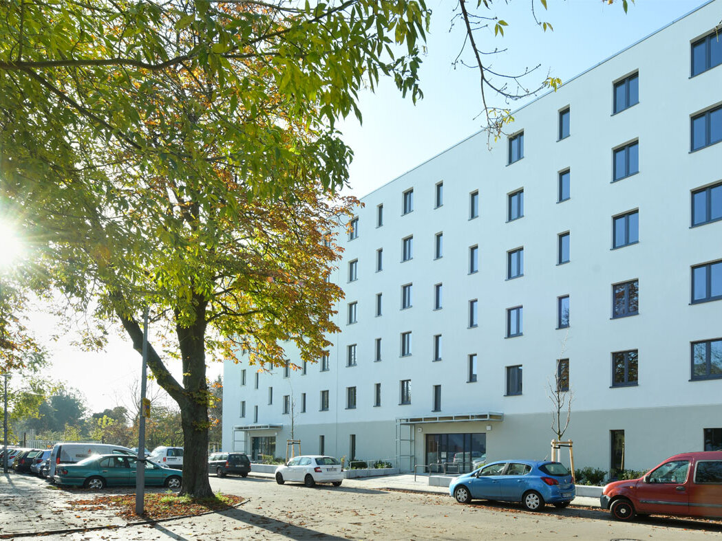 Außenansicht degewo-Neubau Mahlower Straße 2 bis 4 A mit Straße, parkenden Autos und beblätterten Baum links im Vordergrund.