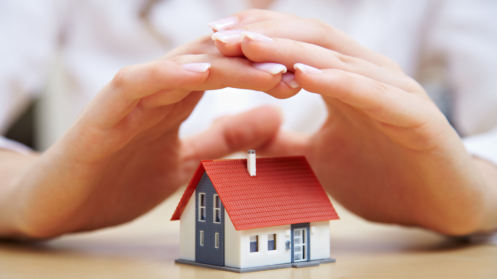 Zwei Hände schützen ein Miniaturhaus auf einer Tischplatte.