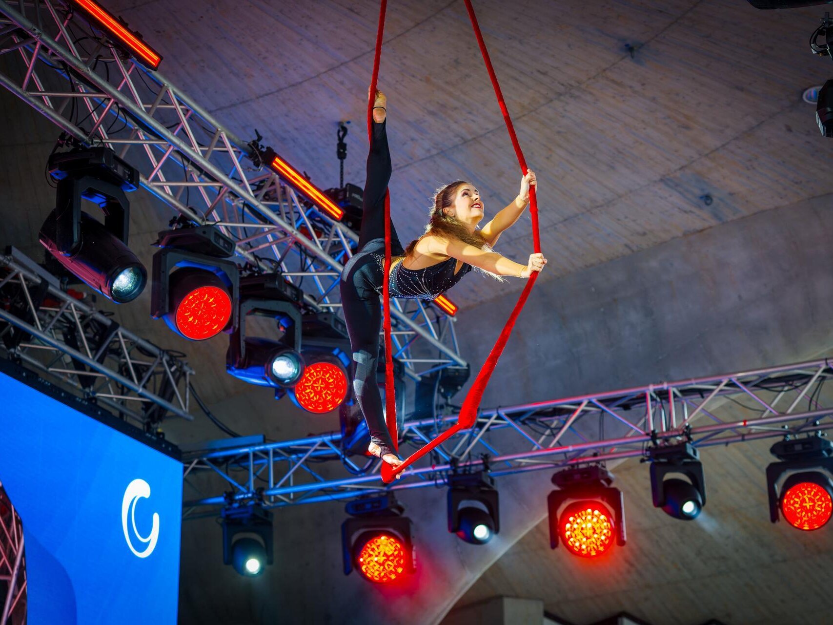 Eine Artistin präsentiert eine Luftakrobatik-Nummer an roten Vertikaltüchern unter dem Bühnendach. Umgeben von farbigen Scheinwerfern führt sie eine anspruchsvolle Pose in mehreren Metern Höhe aus.