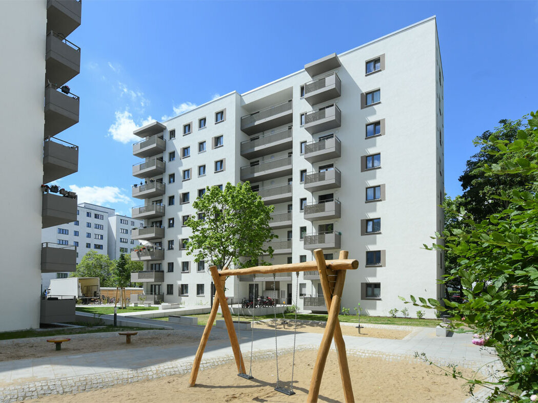 Außenansicht degewo-Neubau Theodor-Loos-Weg 15 und 15 A mit Spielplatz und beblätterten Zweigen rechts im Vordergrund.