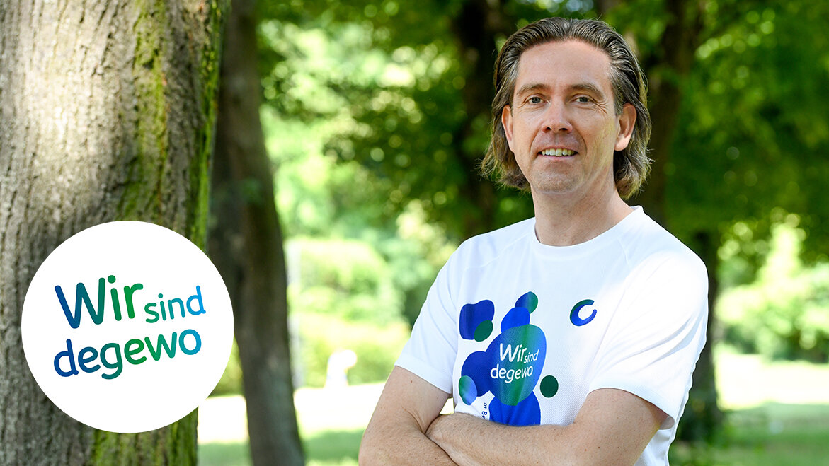 Ein Mann steht in einem Park neben einem Baum. Er trägt ein Sport T-Shirt, seine Arme sind verschränkt.