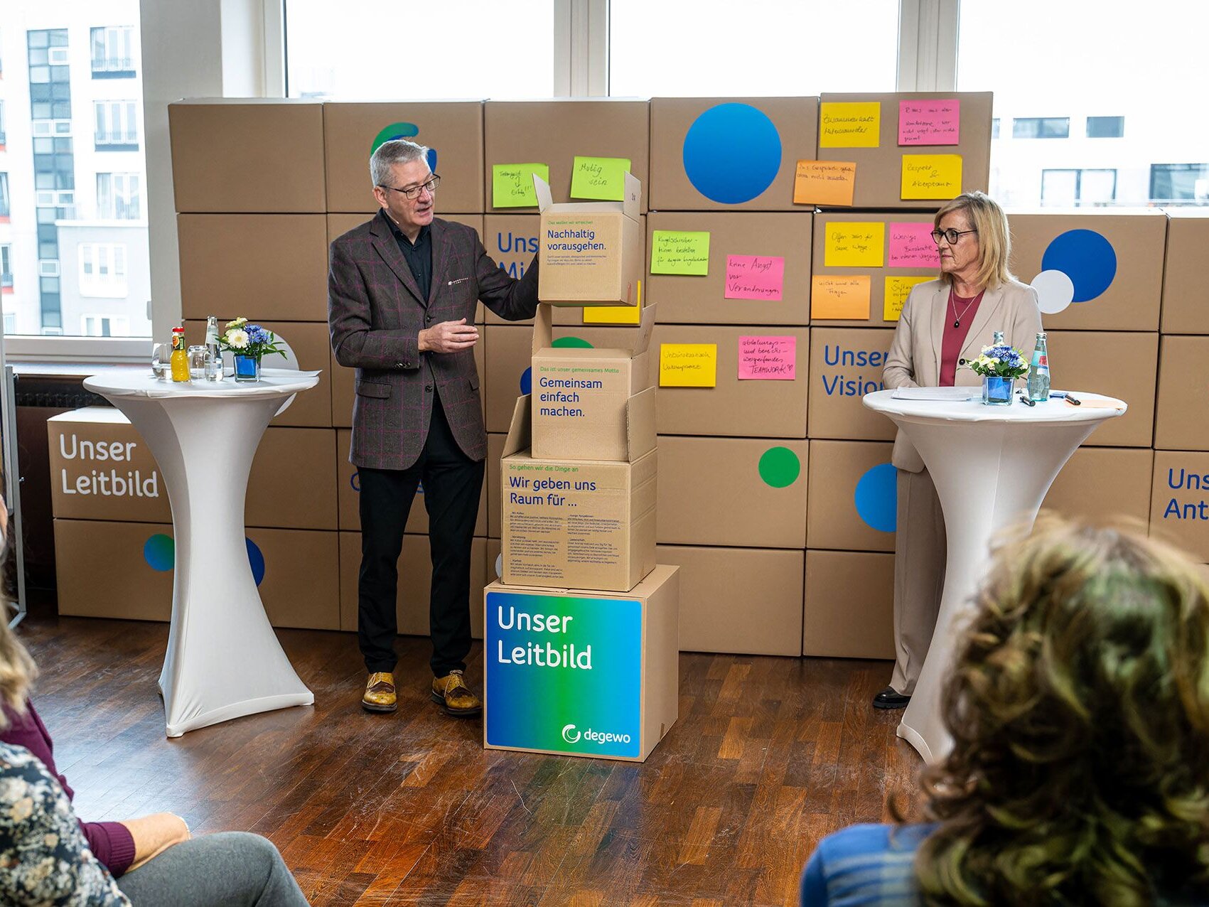 Ein Mann steht neben gestapelten Kartons mit der Aufschrift „Unser Leitbild“ und hält einen weiteren beschrifteten Karton in der Hand. Eine Frau steht ihm gegenüber an einem Stehtisch. Hinter ihnen ist eine Wand aus Kartons mit farbigen Haftnotizen angebracht. Mehrere Personen sitzen im Vordergrund und verfolgen die Präsentation.