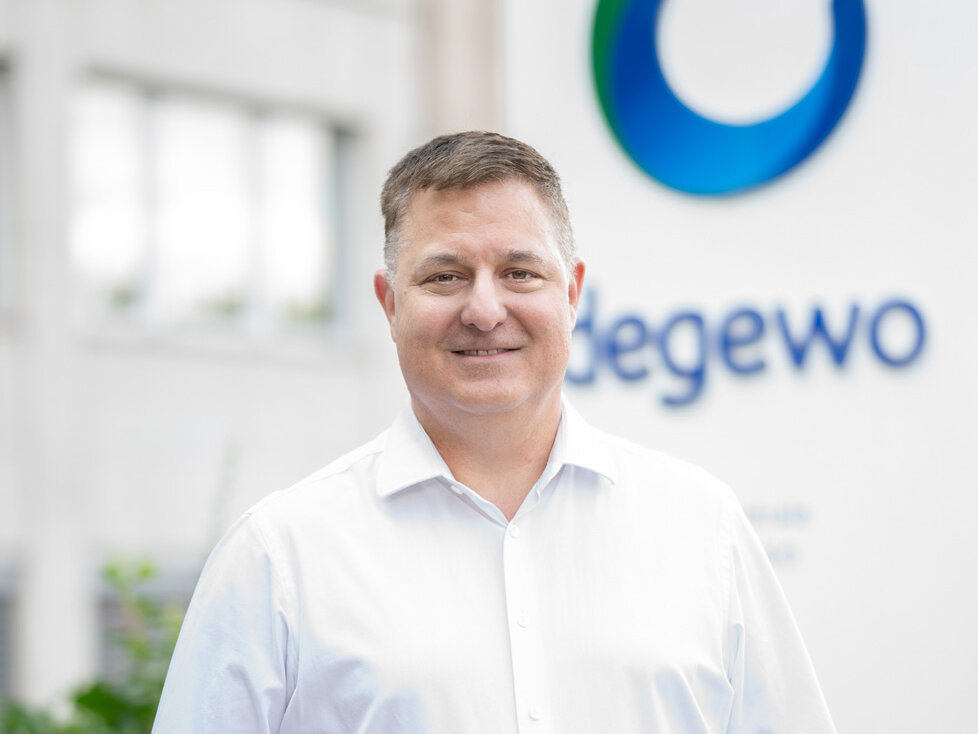 Mann steht im Freien vor einem Gebäude, trägt ein weißes Hemd und lächelt in die Kamera; im unscharfen Hintergrund ist an der Fassade das degewo-Logo zu erkennen.