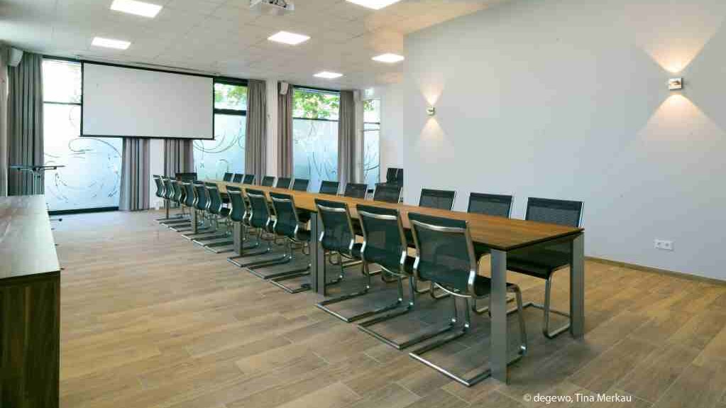 Moderner Konferenzraum mit langem Tisch, mehreren schwarzen Stühlen und einer großen Projektionsleinwand. Helle Fensterfronten sorgen für viel Tageslicht, der Raum ist für Besprechungen und Veranstaltungen ausgestattet.