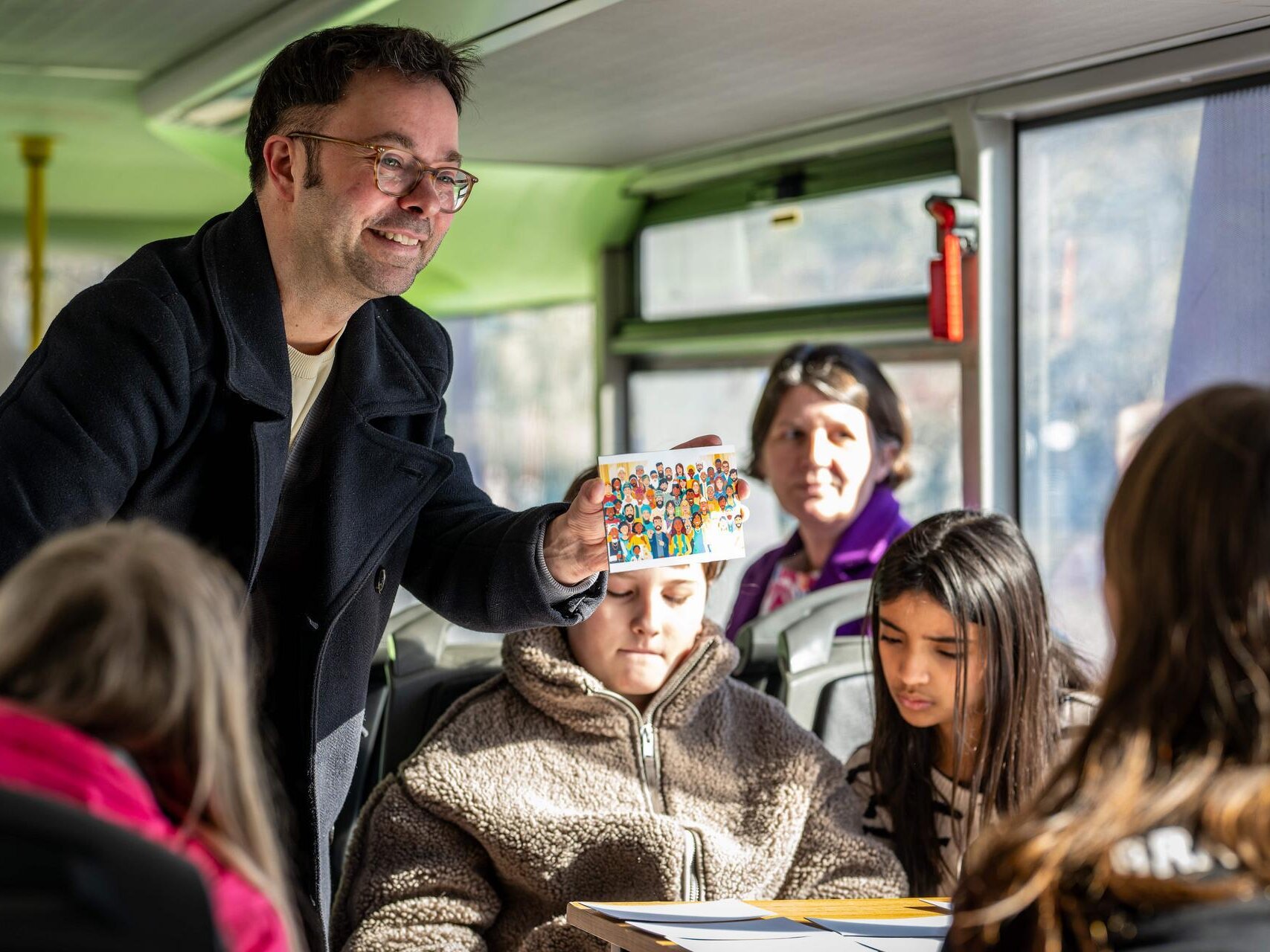 Ein Mann hält eine Karte mit vielen unterschiedlichen Menschen hoch und zeigt sie einem Kind. Im Hintergrund sitzen weitere Kinder und spielen eine Memory-Spiel.