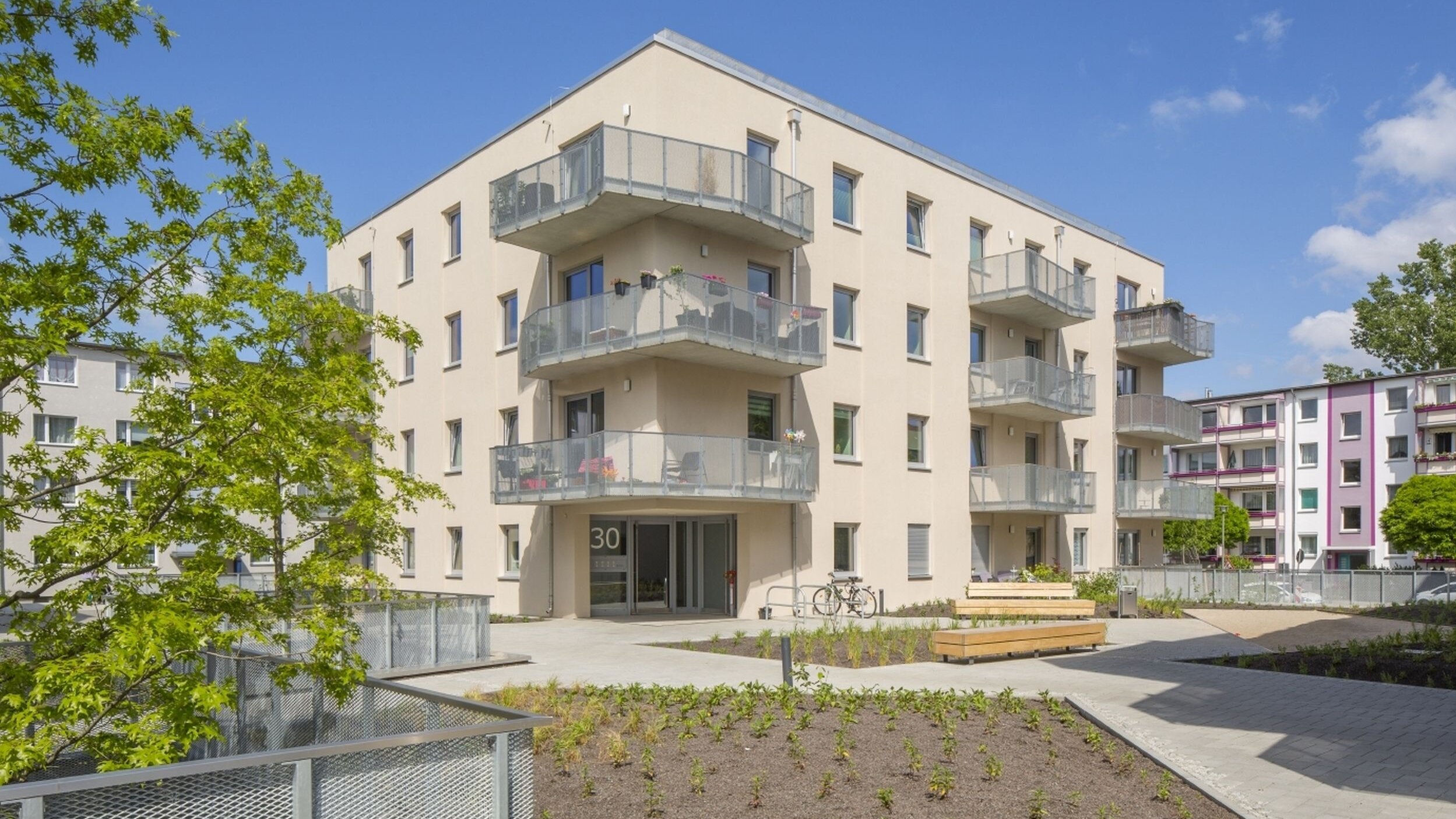 Außenansicht degewo-Neubau Karl-Pokern-Straße 30 mit Bäumen und Zäunen links sowie Beeten, Wegen und Sitzbänken im Vordergrund.