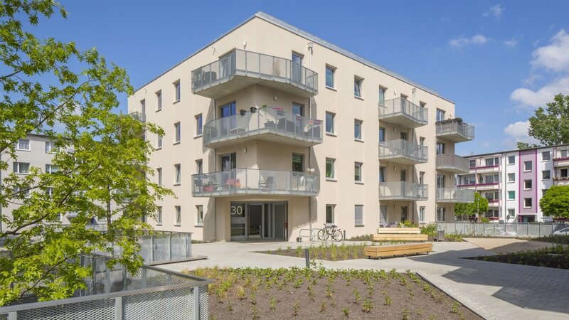 Außenansicht degewo-Neubau Karl-Pokern-Straße 30 mit Bäumen und Zäunen links sowie Beeten, Wegen und Sitzbänken im Vordergrund.