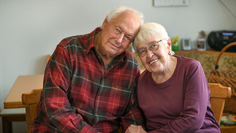 Ein älteres Paar sitzt eng nebenbeinander und schauen lächelnd in die Kamera. Die Köpfe sind aneinander gehalten. 
