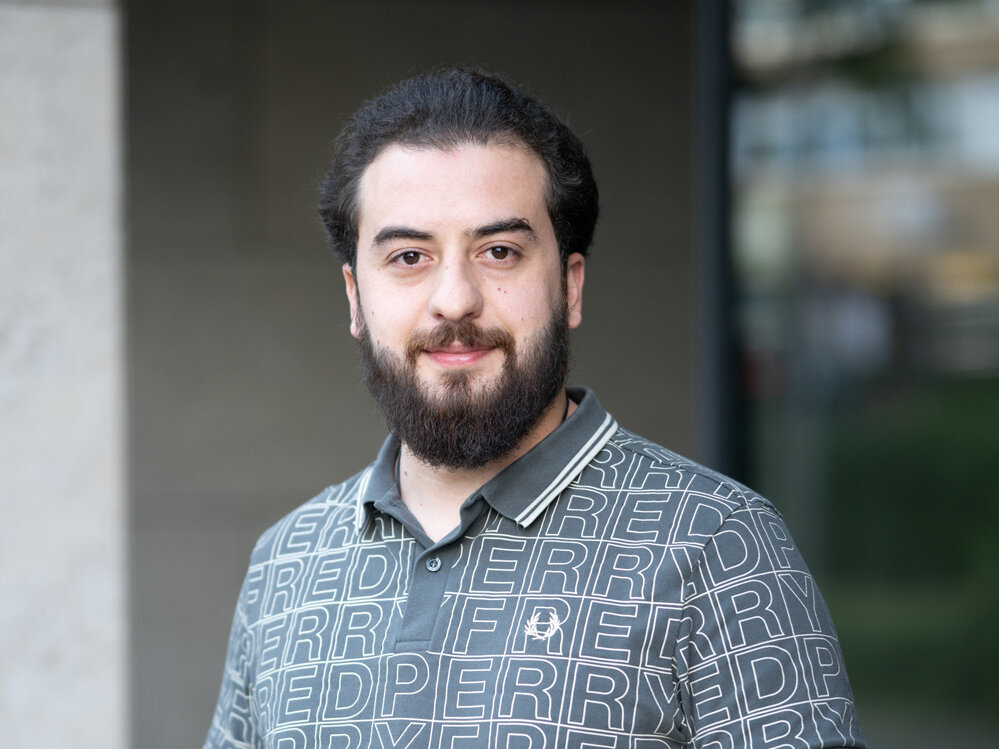 Porträt eines jungen Mannes mit Bart, der in einem graugrünen Poloshirt und hellen Hosen entspannt vor einem Gebäude steht und mit ruhigem Blick in die Kamera schaut.