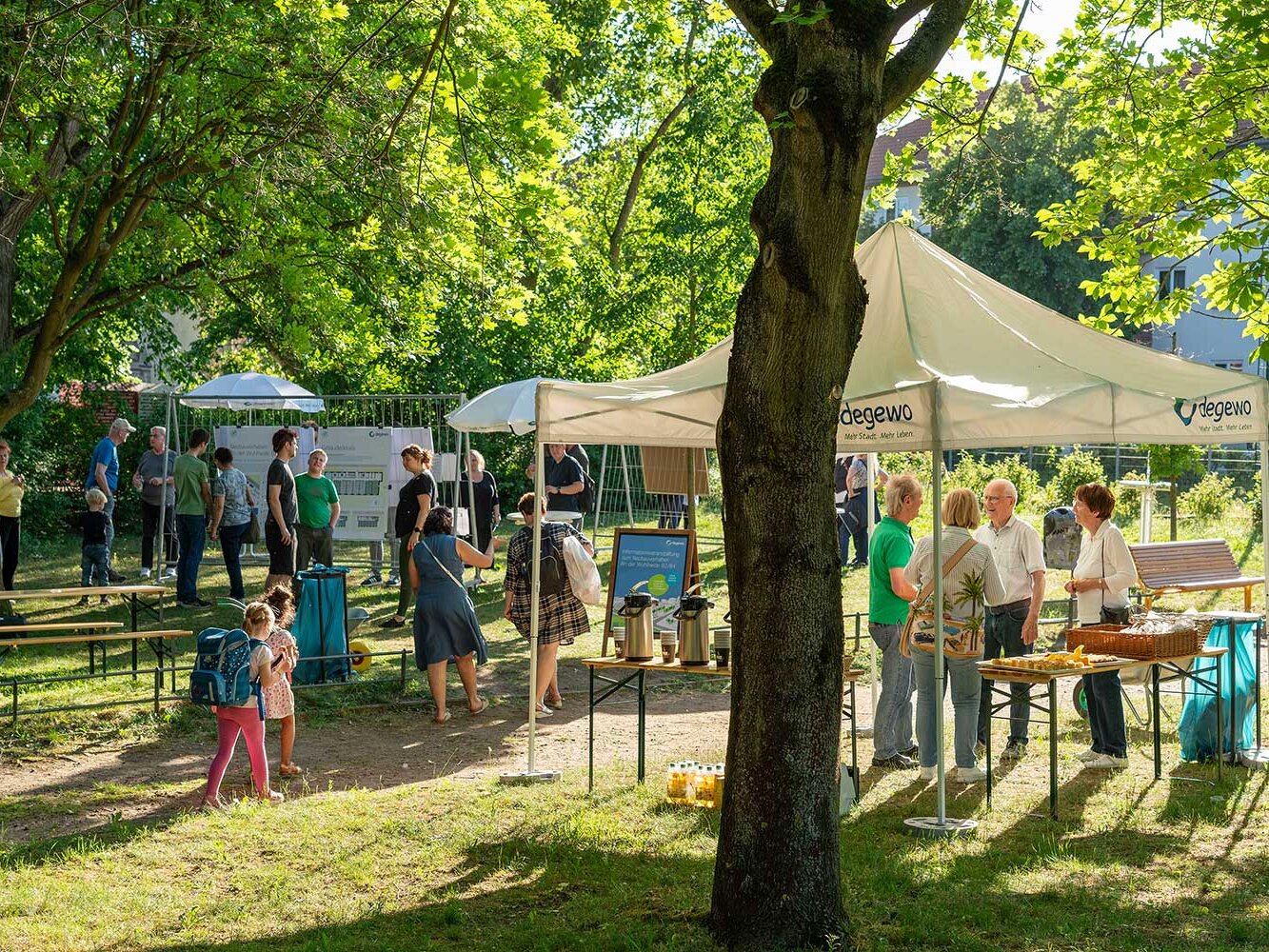 Gesamtansicht der Infoveranstaltung im Park: Besucherinnen und Besucher bewegen sich zwischen Infoständen, Bänken und Tischen. Unter Zelten werden Getränke angeboten, Kinder spielen im Vordergrund.