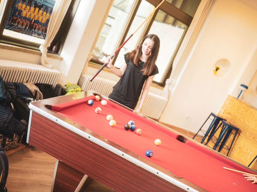 Ein Mädchen spielt an einem Billardtisch und hält einen Queue in der Hand. Der Raum ist hell und mit großen Fenstern ausgestattet.