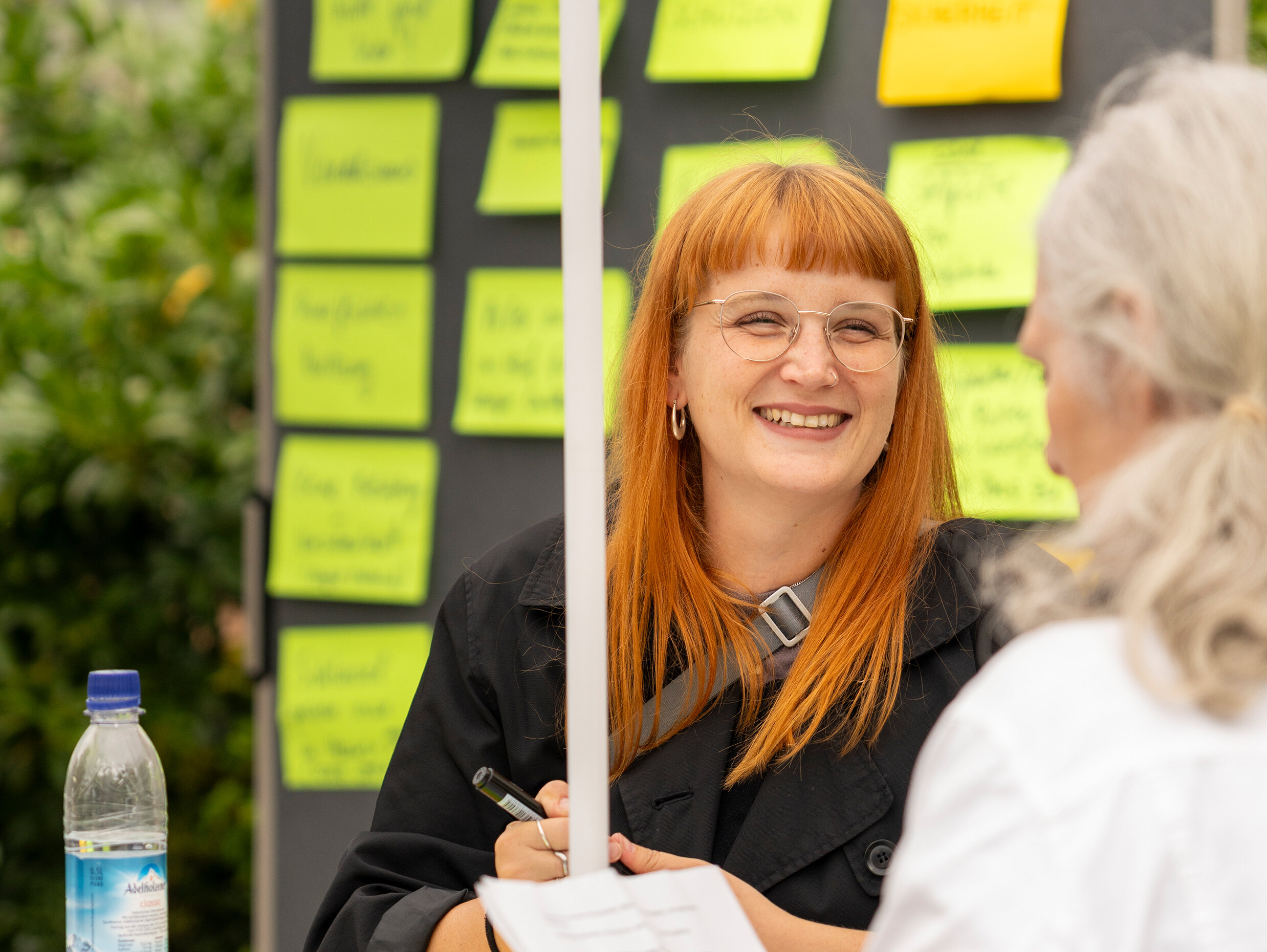 Dialog am Infotisch: Eine Mitarbeiterin im Gespräch mit einer älteren Teilnehmerin, im Hintergrund eine Pinnwand mit gesammelten Anregungen.