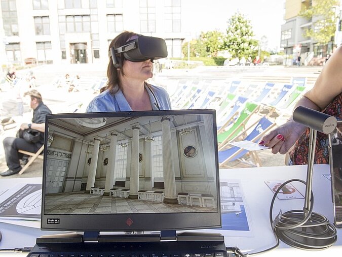 Eine Frau mit einer VR-Brille, davor ein Monitor mit klassischem Gebäude