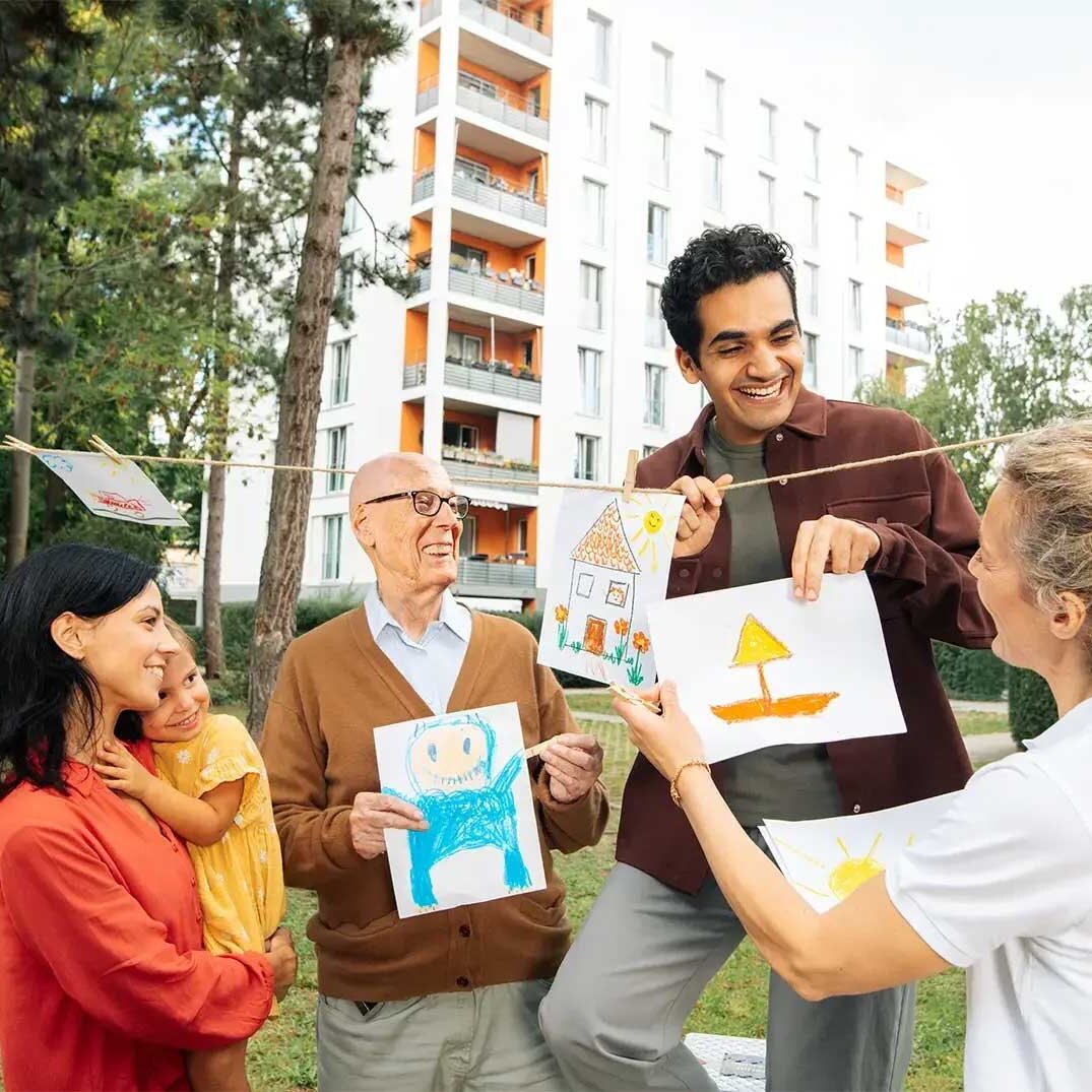 Eine Gruppe Mieterinnen und Mieter schaut sich gemalte Kinderbilder an. Sie sind im Hof und lachen. Im Hintergrund ist ein Mehrfamilienhaus zu sehen.