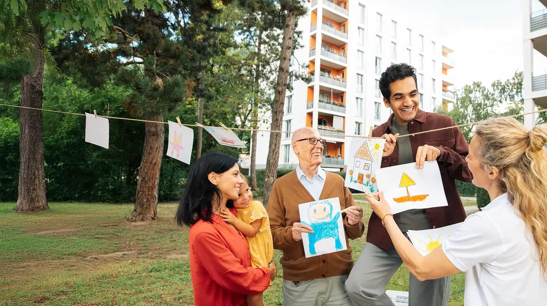 Eine Gruppe Mieterinnen und Mieter schaut sich gemalte Kinderbilder an. Sie sind im Hof und lachen. Im Hintergrund ist ein Mehrfamilienhaus zu sehen.