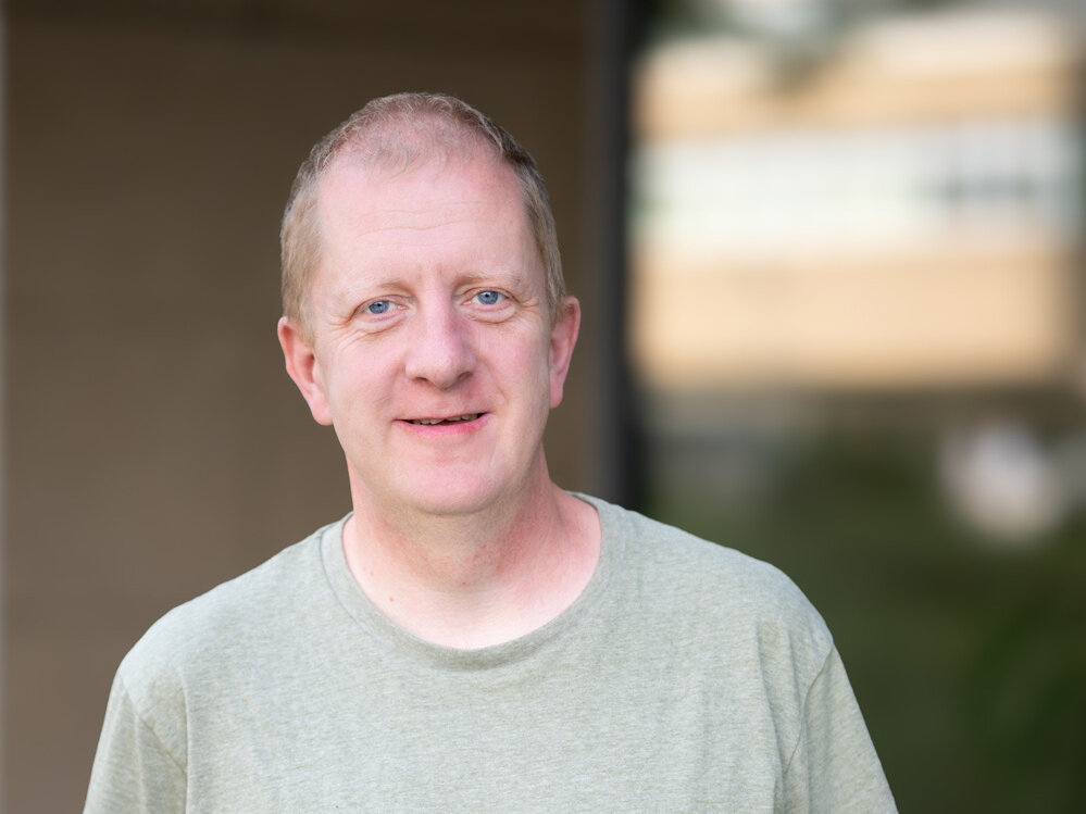 Mann steht im Freien vor einem Gebäude, trägt ein hellgrünes T-Shirt und blickt direkt in die Kamera; der Hintergrund ist weichgezeichnet und zeigt Architektur und Grün.