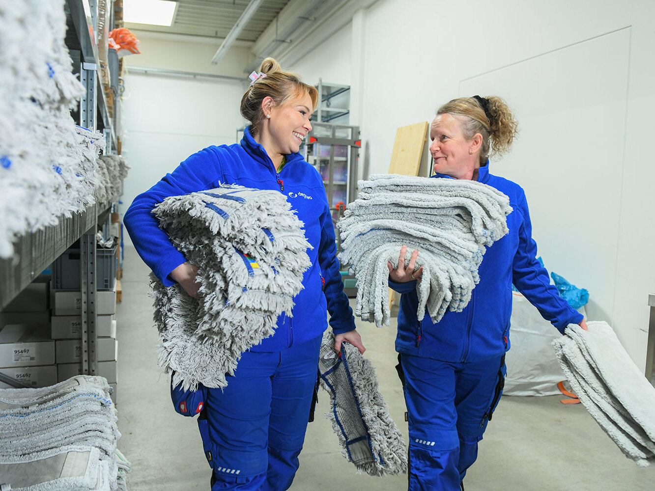 Zwei Mitarbeiterinnen tragen große Stapel gewaschener Mopps durch ein Lager und lächeln sich dabei an.