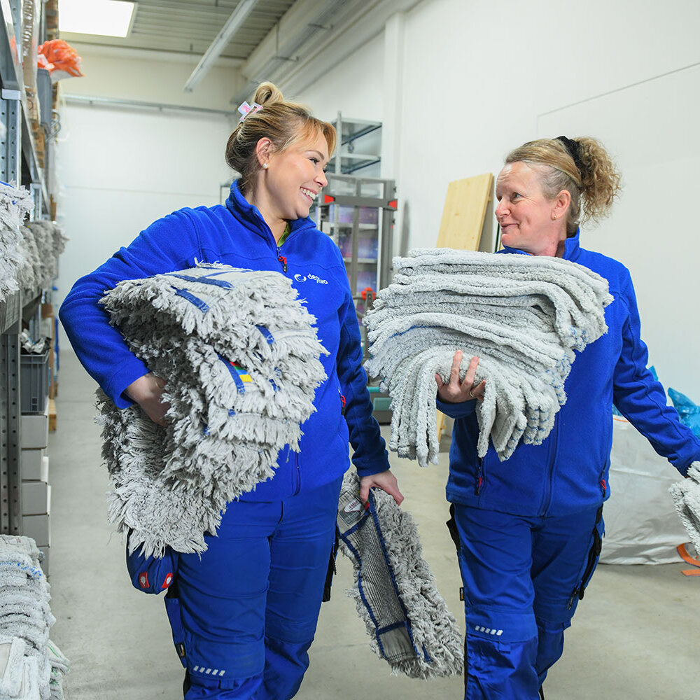 Zwei Mitarbeiterinnen tragen große Stapel gewaschener Mopps durch ein Lager und lächeln sich dabei an.