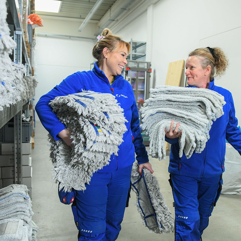 Zwei Mitarbeiterinnen tragen große Stapel gewaschener Mopps durch ein Lager und lächeln sich dabei an.