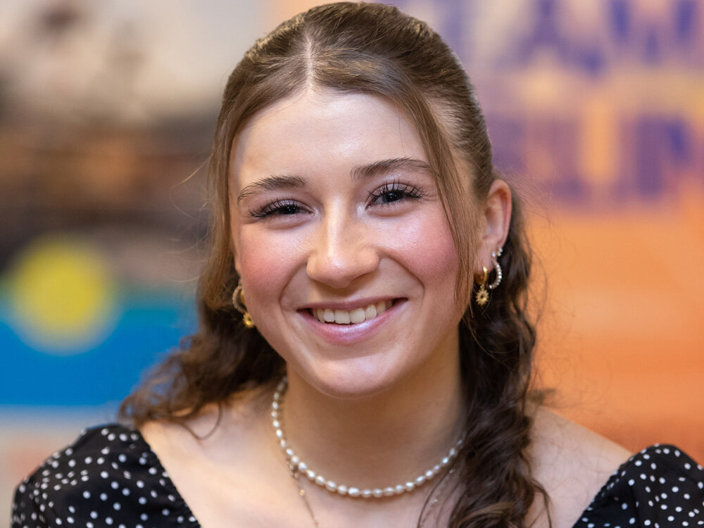 Junge Frau mit gepunkteter Bluse und Perlenkette lächelt in die Kamera. Der Hintergrund zeigt verschwommene Sponsorenlogos.