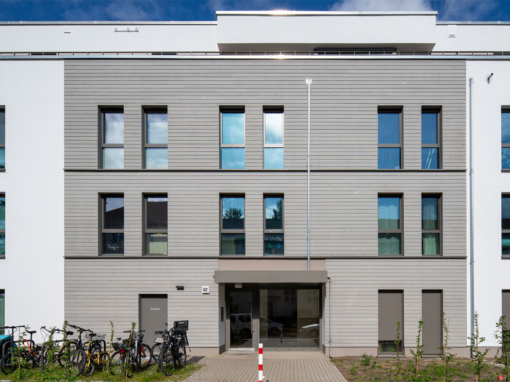 Außenansicht degewo-Neubau Stillerzeile 62: Eingangsbereich mit Fahrradständern links und Beeten rechts vor dem Gebäude.