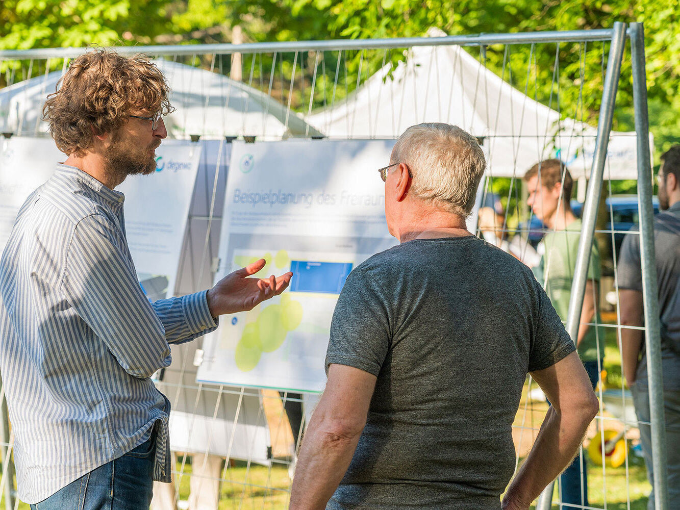Zwei Männer diskutieren vor einer Tafel mit einer Beispielplanung für den Freiraum. Einer erklärt gestikulierend Details, im Hintergrund sind weitere Besucherinnen und Besucher zu sehen.
