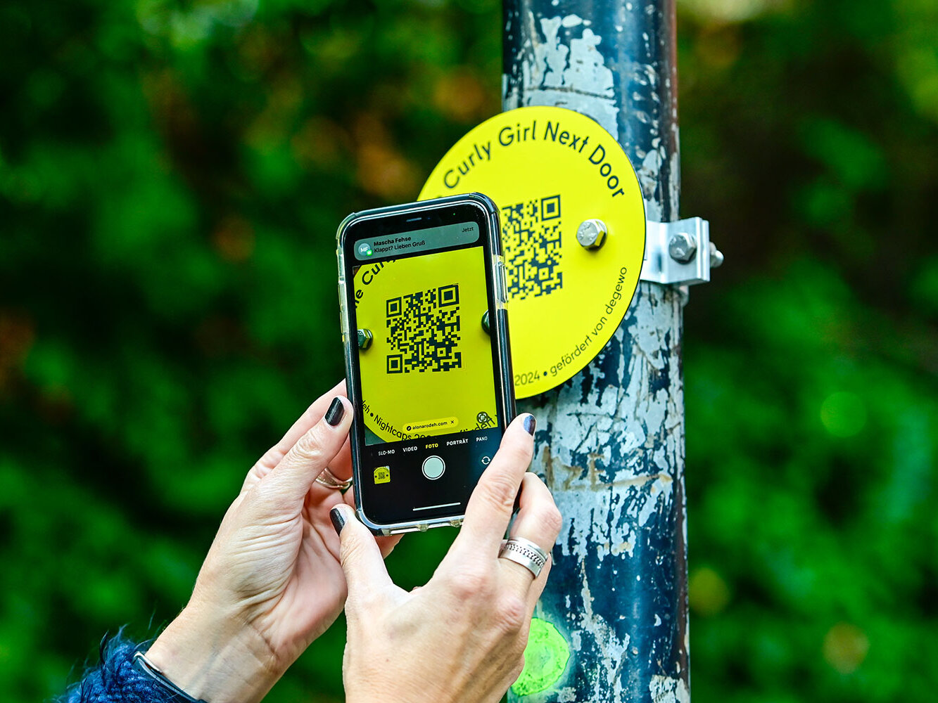 Eine Person scannt mit dem Smartphone einen gelben QR-Code an einer Straßenleuchte. Die Nahaufnahme zeigt die Hände und den QR-Code gut lesbar.