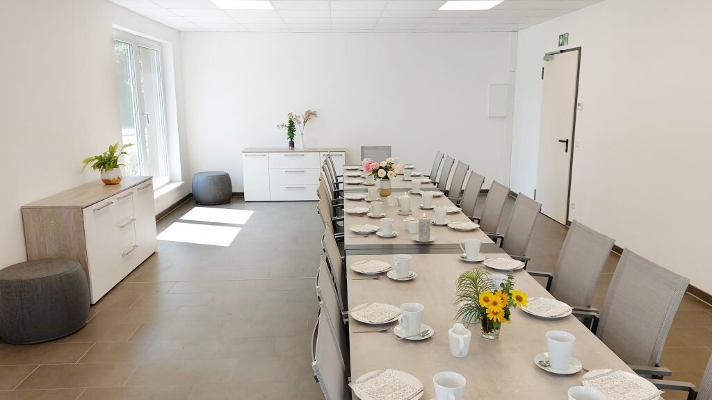 Heller Veranstaltungsraum mit einer langen, festlich gedeckten Tafel für mehrere Personen. Auf den Tischen stehen Tassen, Teller und kleine Blumenvasen, entlang der Wände befinden sich Schränke und Sitzhocker.