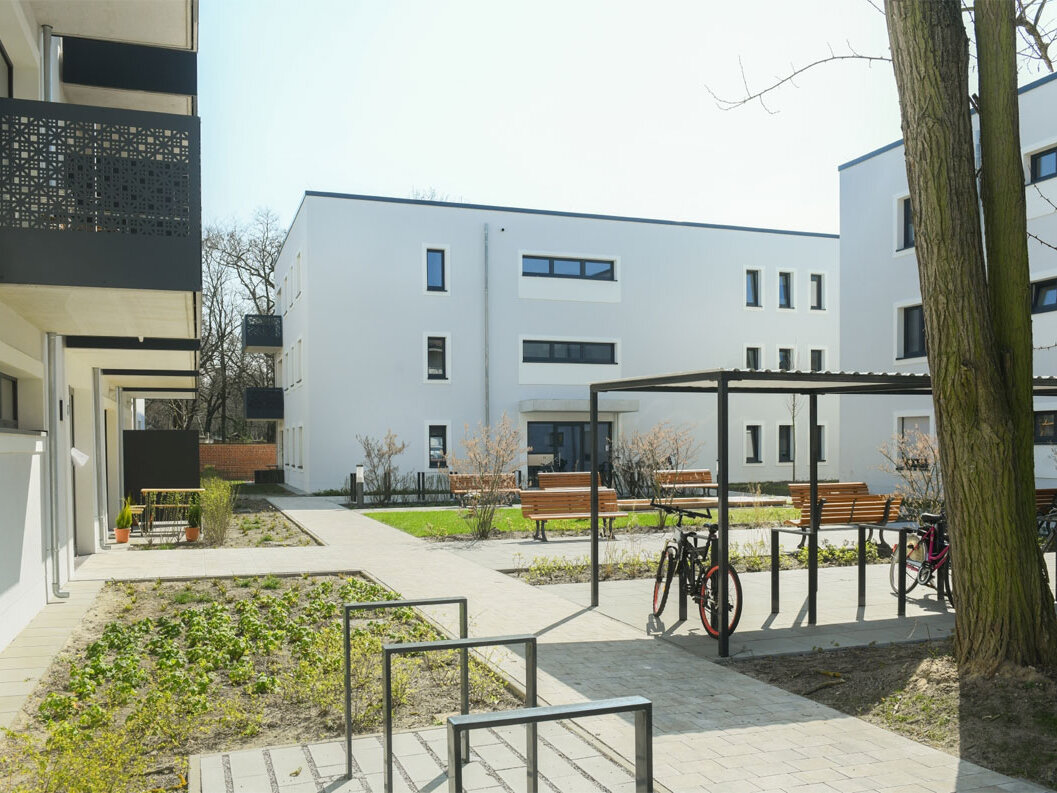Außenansicht degewo-Neubau Rudower Straße 47 bis 51: Hofseite mit Beeten, teils überdachten Fahrradständern und Sitzbänken sowie rechts im Vordergrund zwei Baumstämmen.
