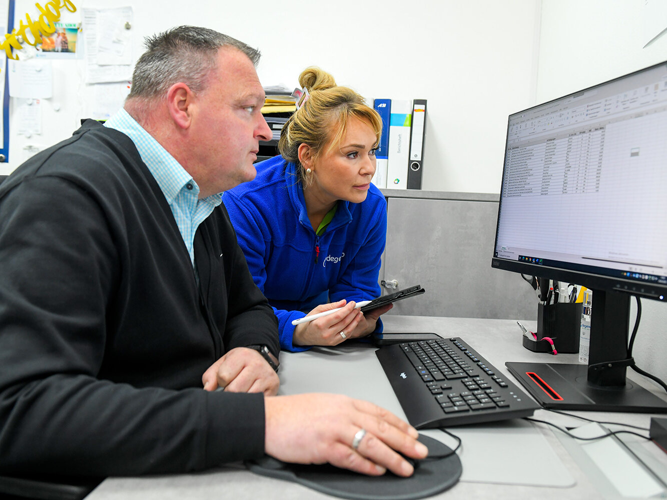 Zwei Mitarbeitende sitzen vor einem Computer und betrachten gemeinsam eine Tabellenkalkulation während eine der Personen Notizen auf einem Tablet macht.