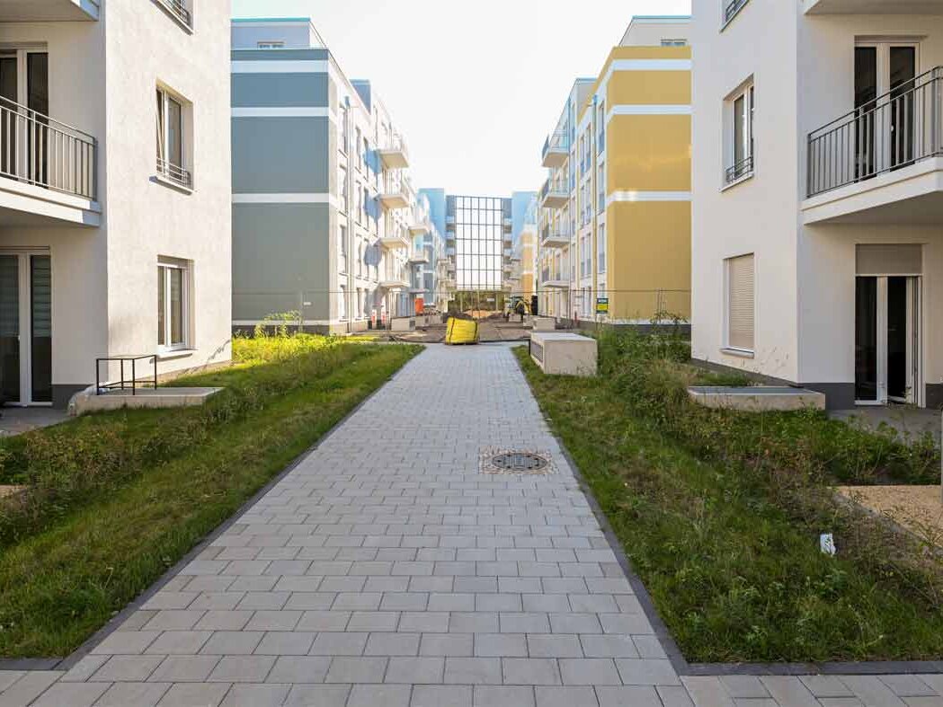 Blick entlang eines gepflasterten Weges zwischen mehreren Neubauten am Eisenhutweg. Die Wohngebäude mit hellen Fassaden und farbigen Akzenten rahmen einen begrünten Innenbereich. Balkone und Erdgeschosszugänge sind sichtbar und vermitteln die Struktur des neu entstandenen Quartiers.