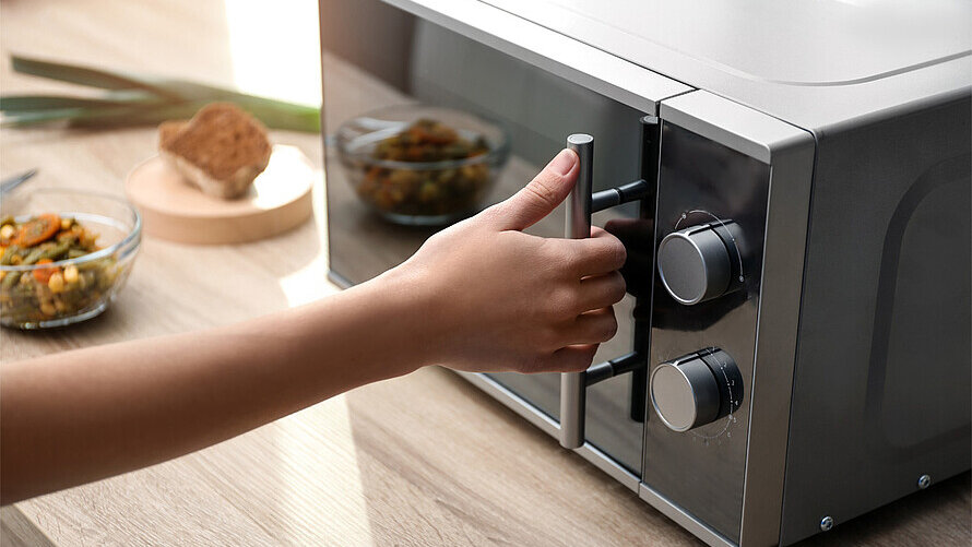 Eine Person greift mit dem rechten Arm nach dem Griff einer Mikrowelle.