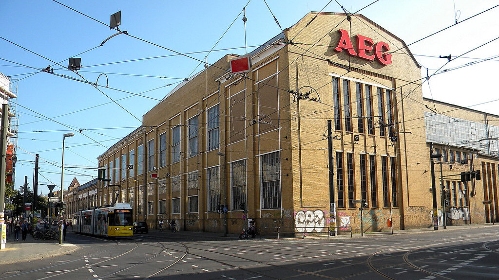 Ein großes historisches Industriegebäude mit AEG-Schriftzug steht an einer Straßenecke. Davor laufen Oberleitungen und eine Straßenbahn über die Straße.