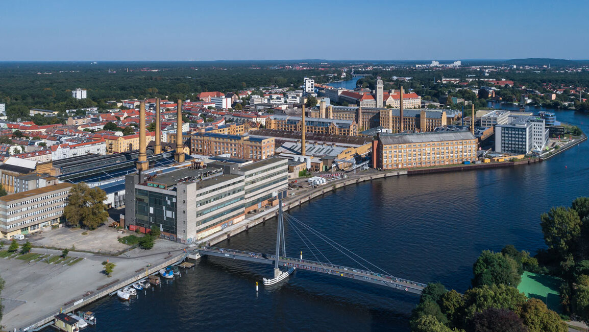 Ein Luftbild zeigt einen Teil des Berliner Stadtteils Oberschöneweide mit einer Brücke, Industriegebäuden, Wohnhäusern, einem Fluss und Grünflächen.